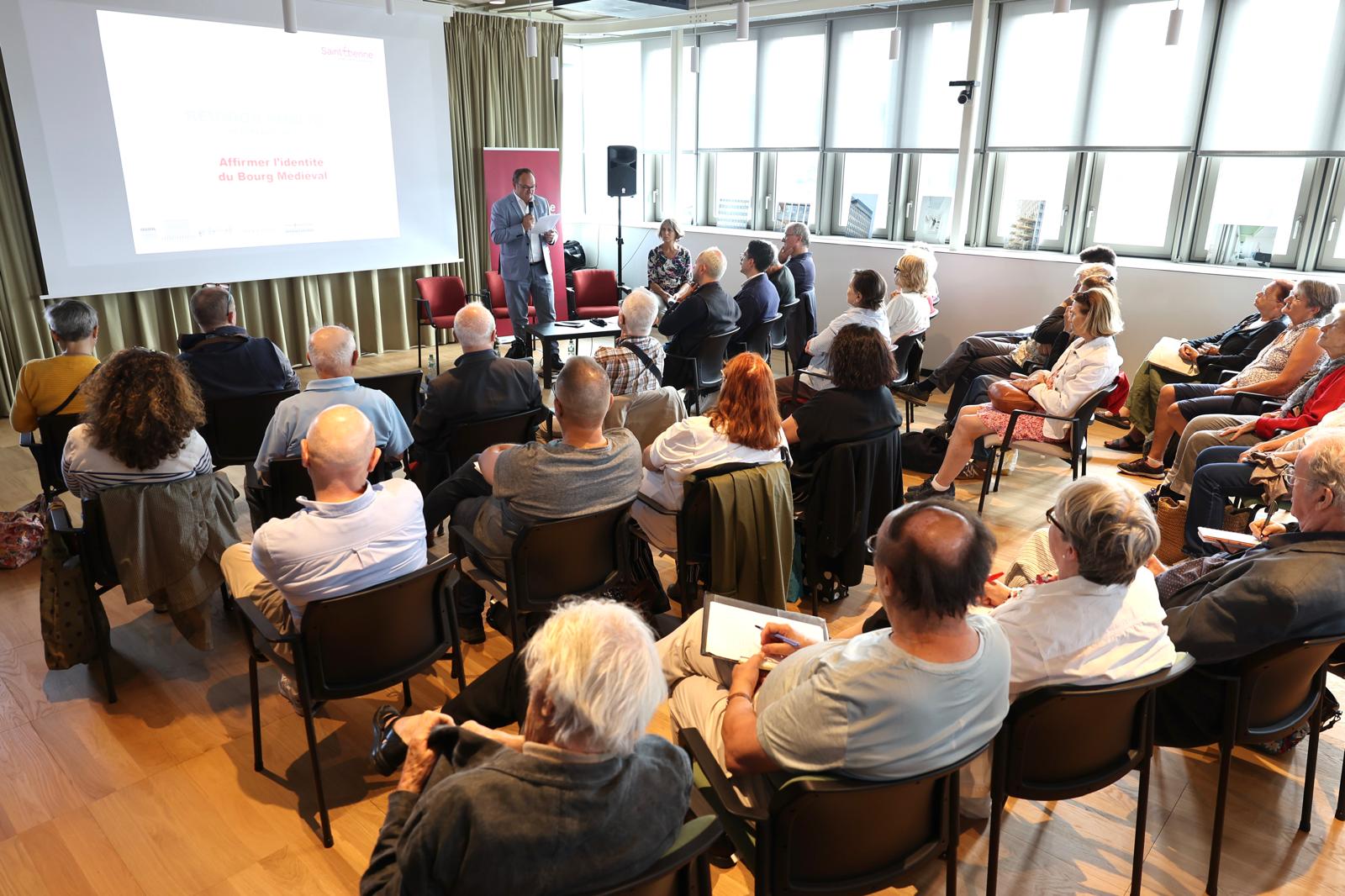 réunion avec un groupe de personnes assises et un personne avec un micro à côté un écran qui projette du conte