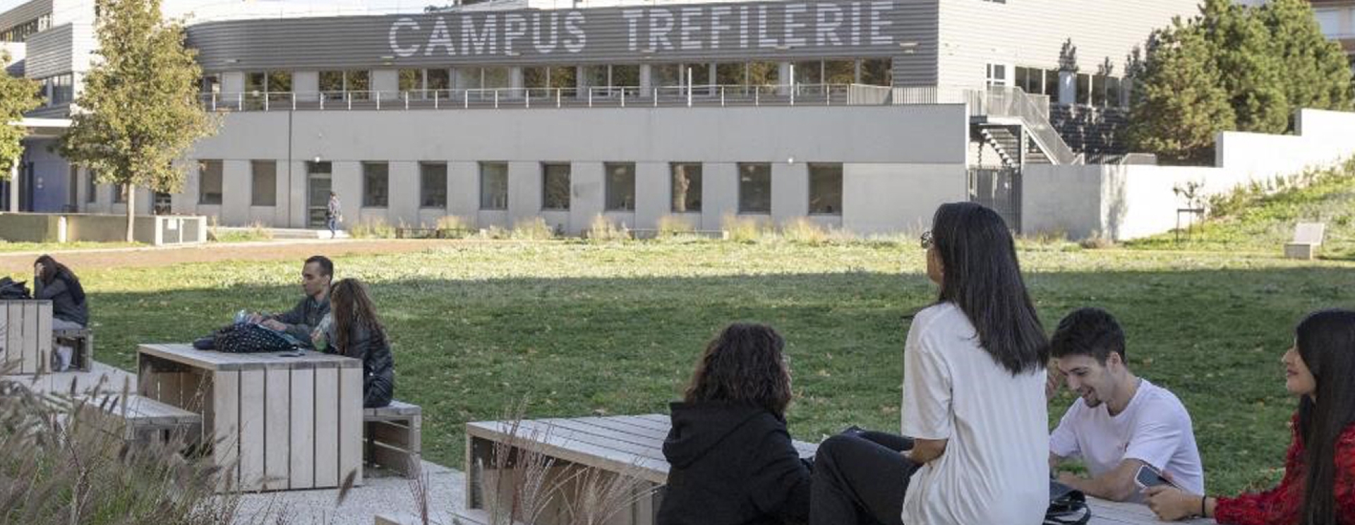 Des jeunes assis à une table, une grande pelouse et un bâtiment avec la mention "Campus Tréfilerie"