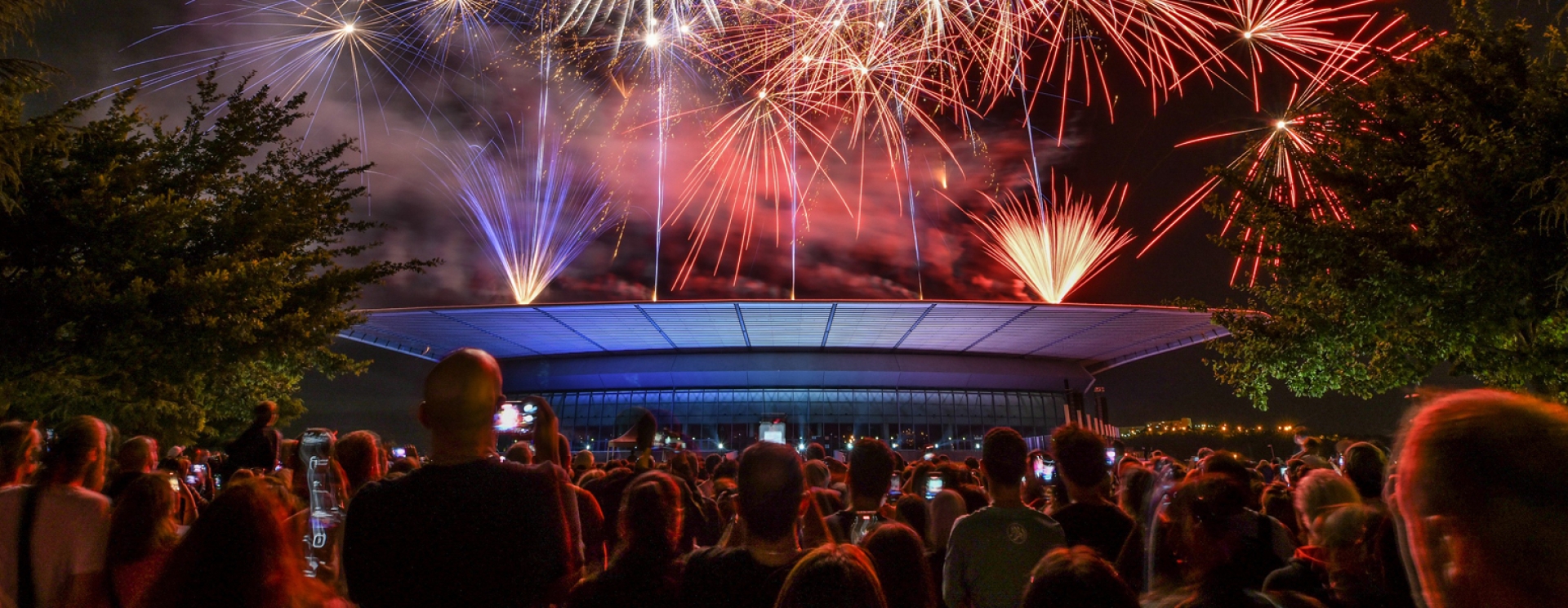 une foule devant un bâtiment surplombée d'un feu d'artifice