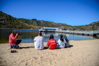 une personne qui filme des jeunes sur une plage, en arrière plan un bateau croisière