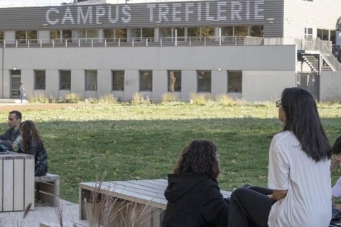 Des jeunes assis à une table, une grande pelouse et un bâtiment avec la mention "Campus Tréfilerie"